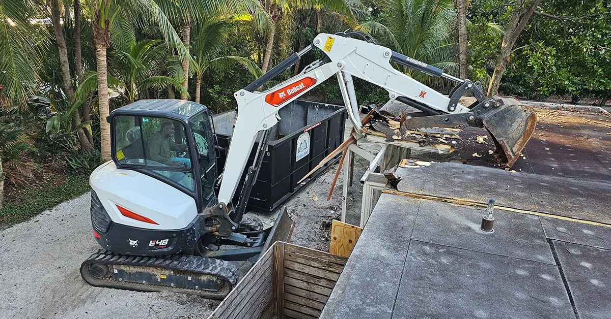 The Smart Way to Handle Waste: Versatile Dumpster Rentals in Southwest Florida. Featuring a Bobcat backhoe demolishing an old and damaged home in Naples and neatly disposing of the debris in the nearby compact dumpster.