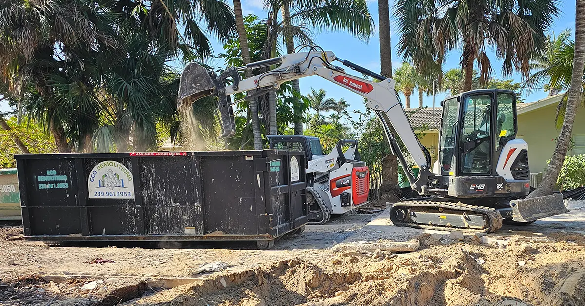 The best dumpster rentals available in Southwest Florida, delivered by Eco Demolitions. Featuring a Bobcat backhoe dumping a bucket-load of residential demolition debris into a nearby compact dumpster.