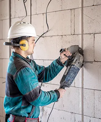 Worker wearing a green and black jacket cutting through old cinder blocks with a specialized circular saw.