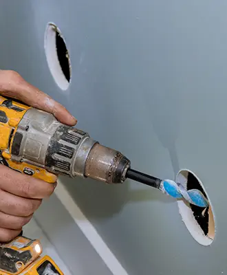 Photo of a person drilling into a stud through a utility hole in the drywall to add a supplementary pathway for wiring.