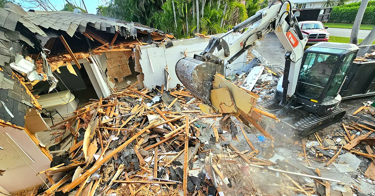 A Bobcat backhoe picking up debris from the corner of a house in its arm.