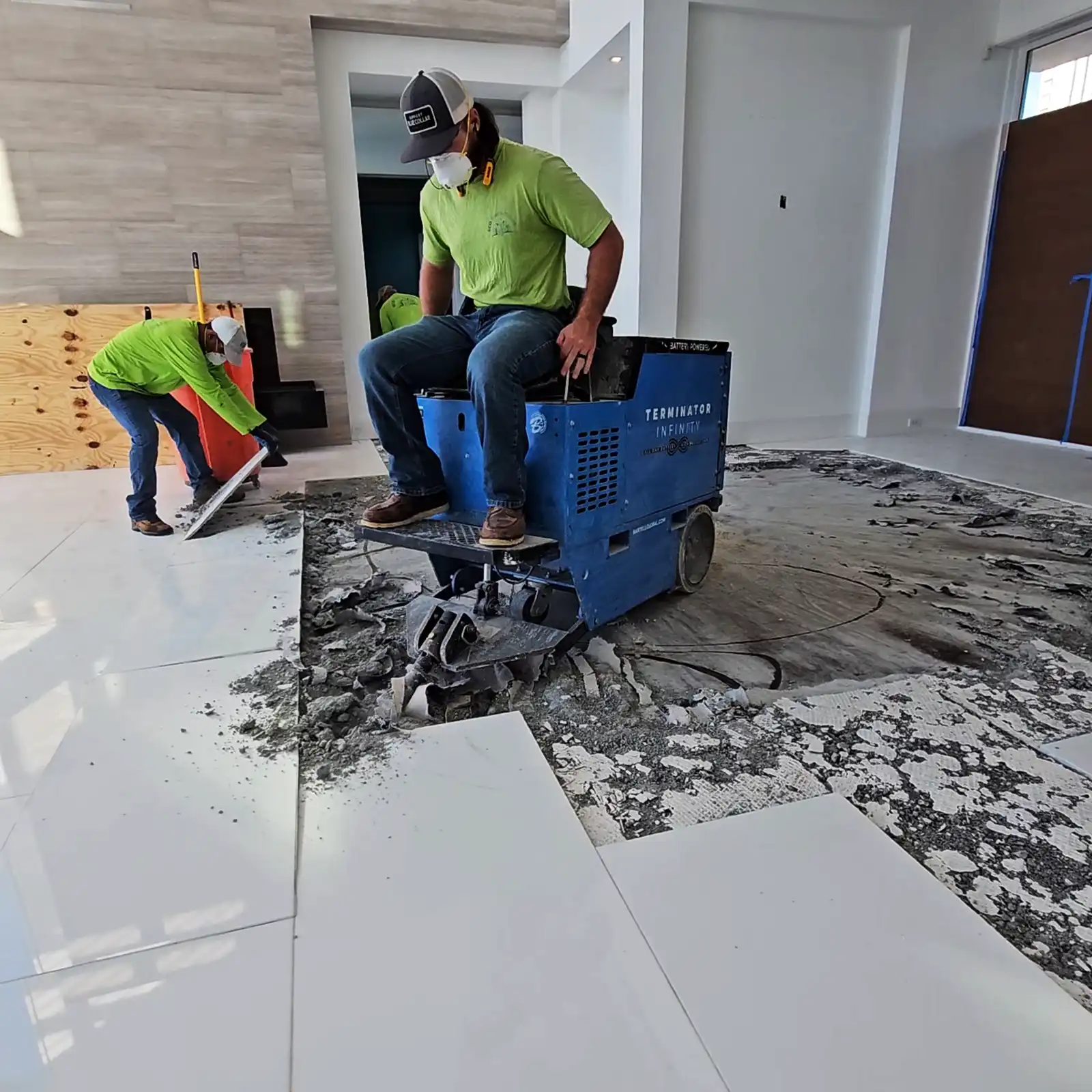 The Importance of Professional Floor Removal in Renovations Projects. Featuring an Eco Demolitions employee riding an industrial floor scraper to break up the white tile in a Naples condo while another employee lifts and removes a whole tile behind him.