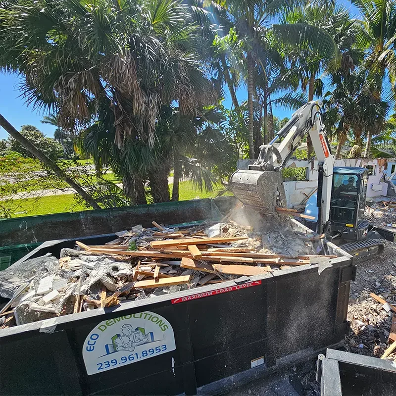 A Bobcat backhoe filling a compact dumpster at an Eco Demolitions job site with construction debris.